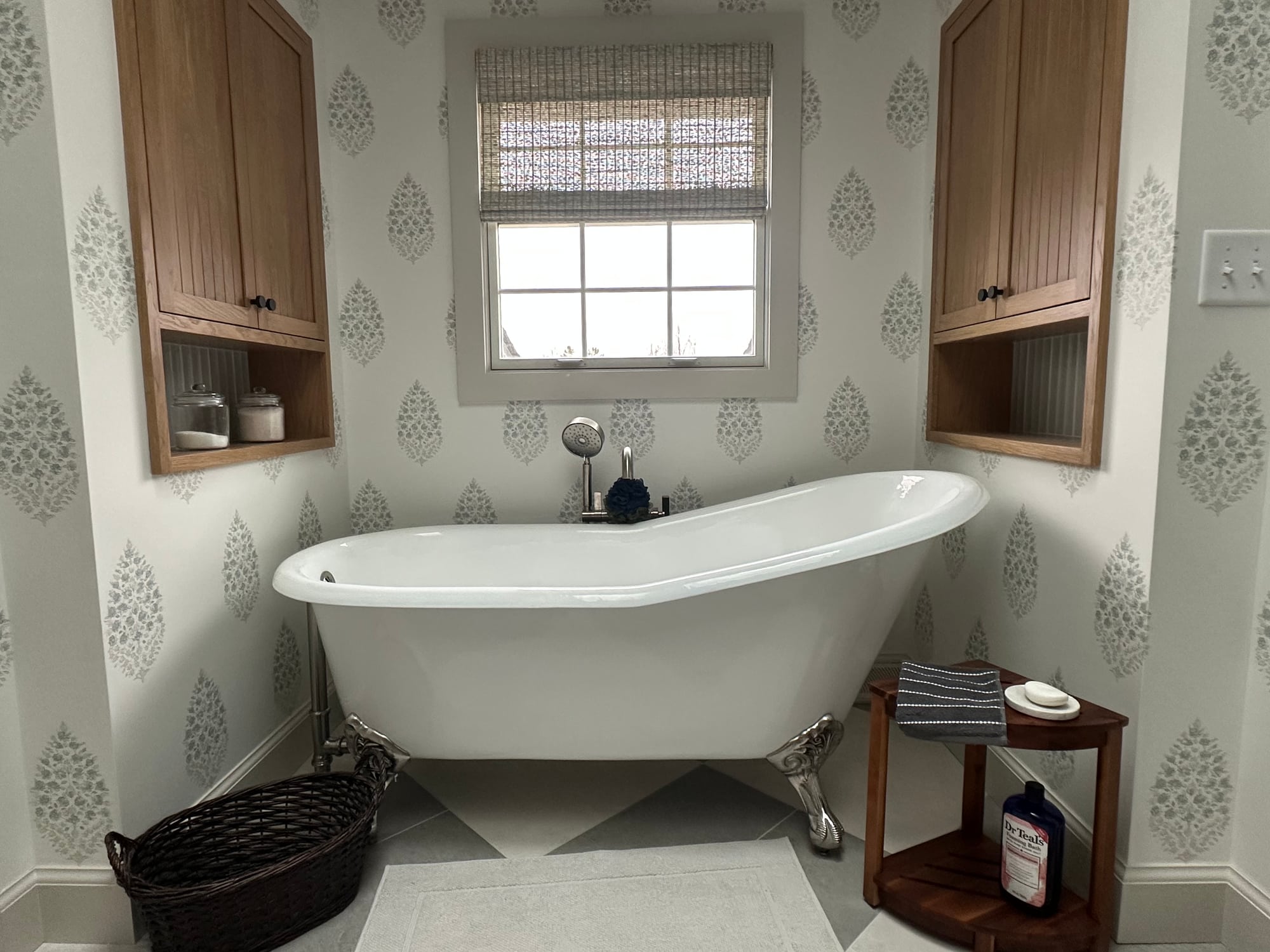 A bath with a freestanding clawfoot tub, patterned wallpaper, wood cabinetry, and a woven shade on the window