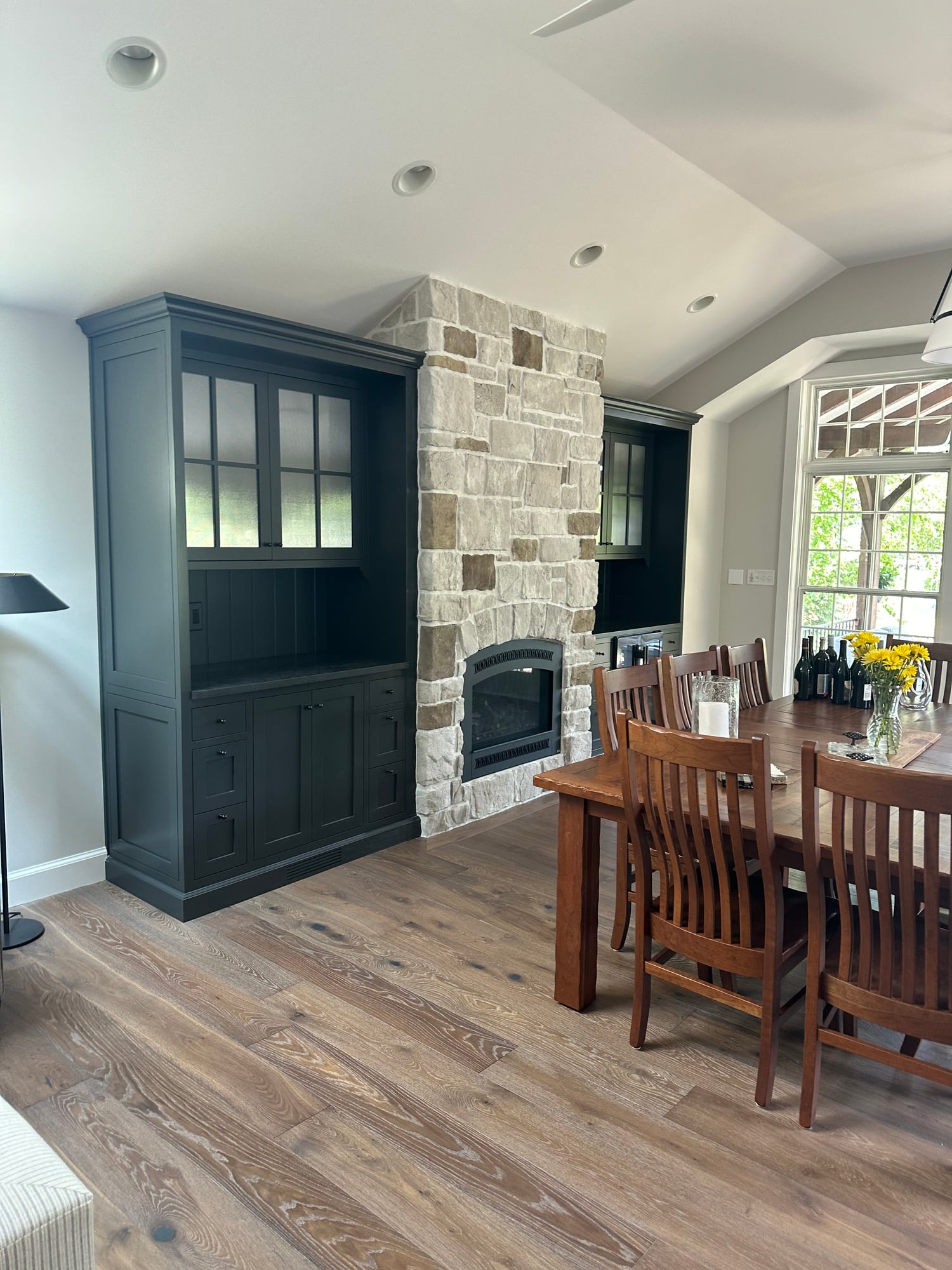 A dining room with a deep green built-in hutch beside a stone fireplace and a wood dining table set by tall windows