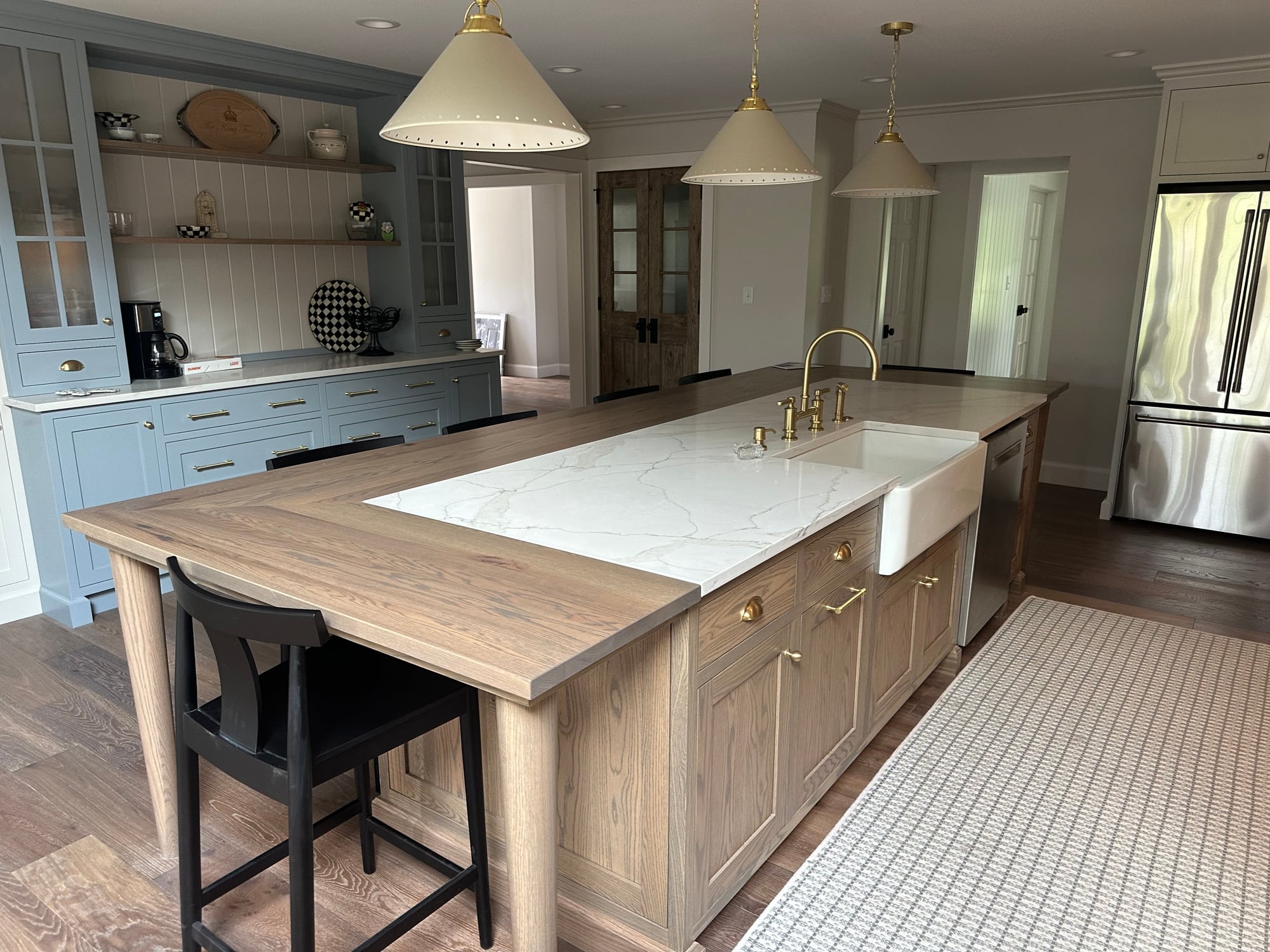 A custom kitchen with a long wood-topped island, farmhouse sink, brass pendant lights, and blue-gray cabinetry