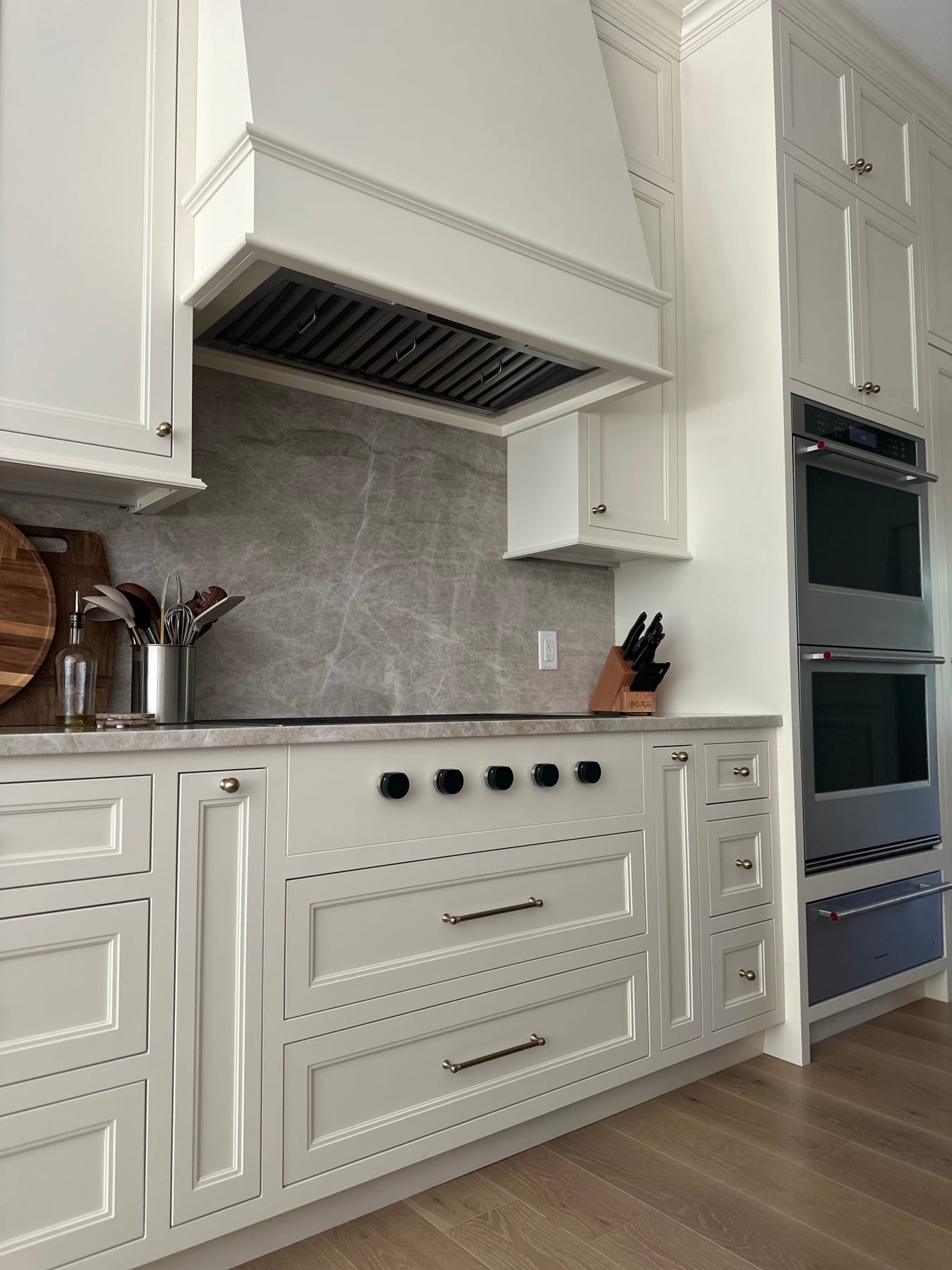 A white kitchen run with a stone-slab backsplash, professional hood, and double wall ovens