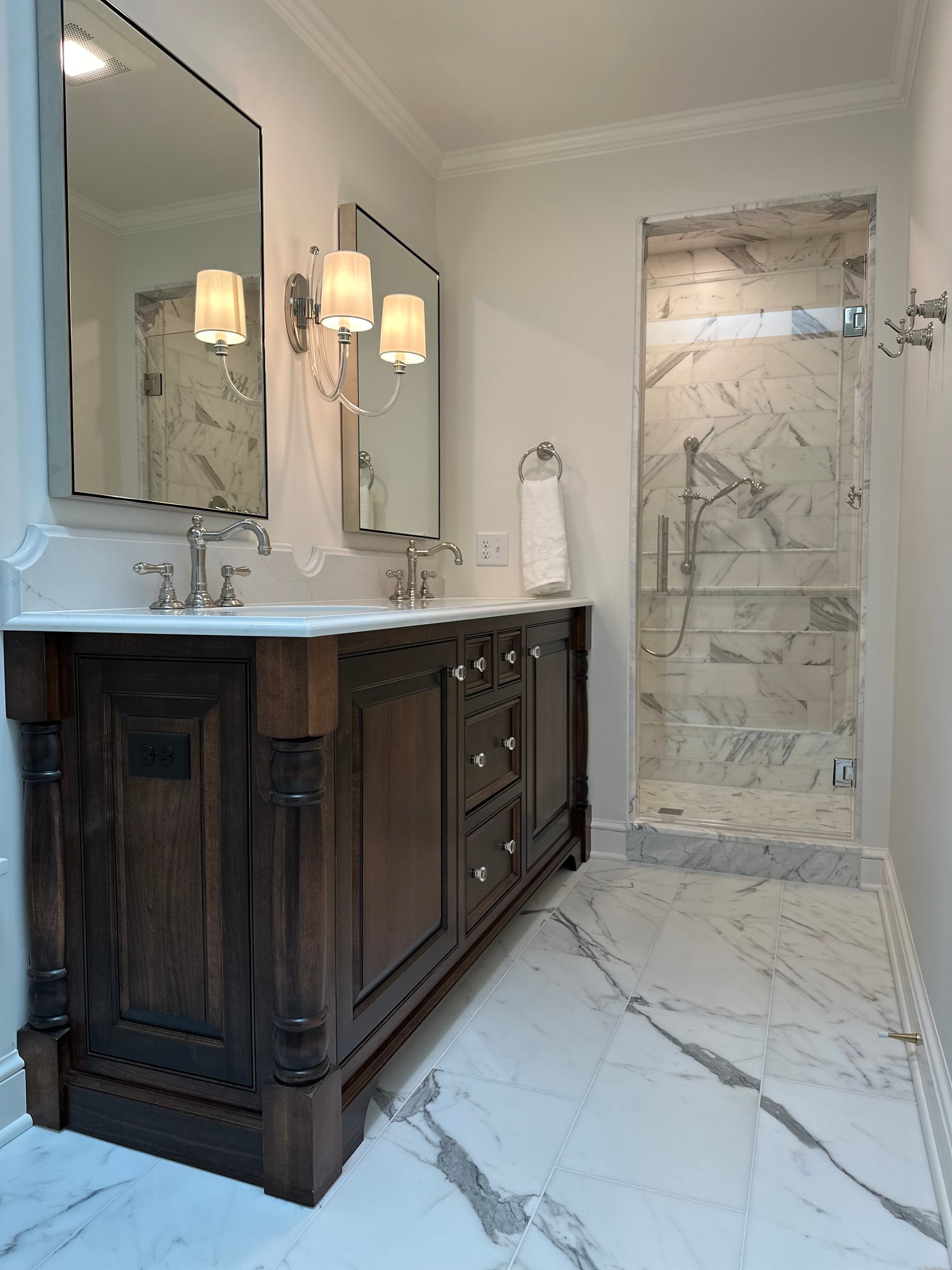 A marble bath with a dark turned-leg double vanity, twin mirrors with sconces, and a glass-door marble shower beyond