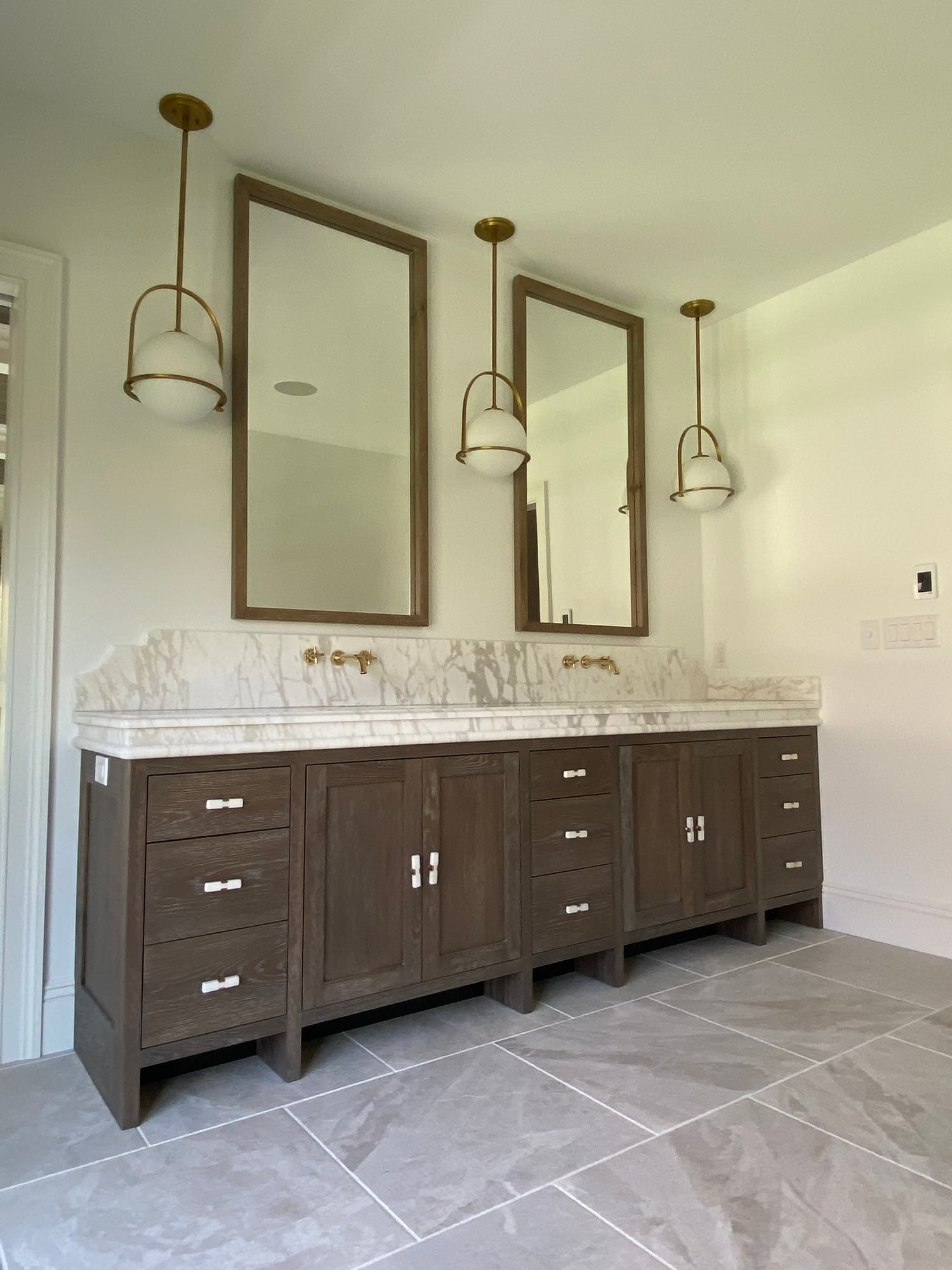 A primary bath double vanity in stained oak with a tiered marble top, twin framed mirrors, and brass pendant lights